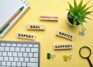 Flat lay of office desk with wooden blocks connected by strings labeled 'Consulting,' 'Goal,' 'Expert,' 'Advice,' and 'Support,' symbolizing business strategy and teamwork. Surrounding items include a computer keyboard, pen, binder clips, magnifying glass, and a potted plant on a yellow background.