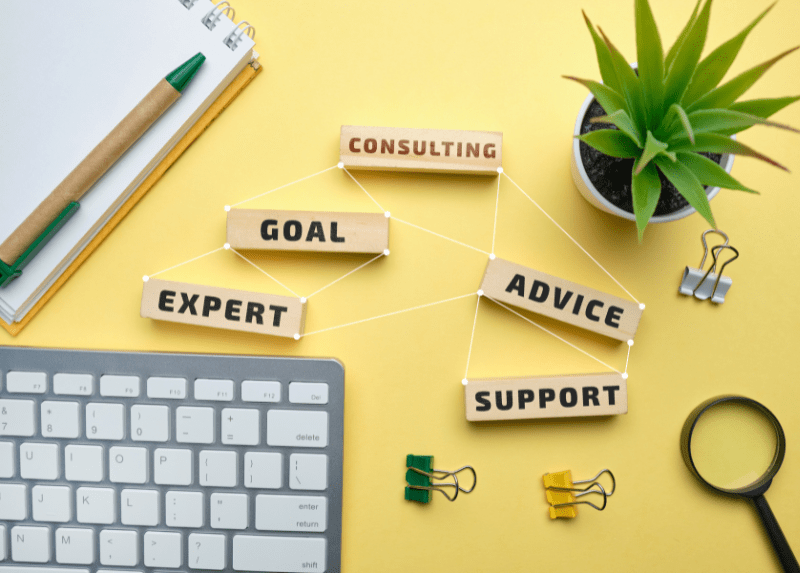 Flat lay of office desk with wooden blocks connected by strings labeled 'Consulting,' 'Goal,' 'Expert,' 'Advice,' and 'Support,' symbolizing business strategy and teamwork. Surrounding items include a computer keyboard, pen, binder clips, magnifying glass, and a potted plant on a yellow background.