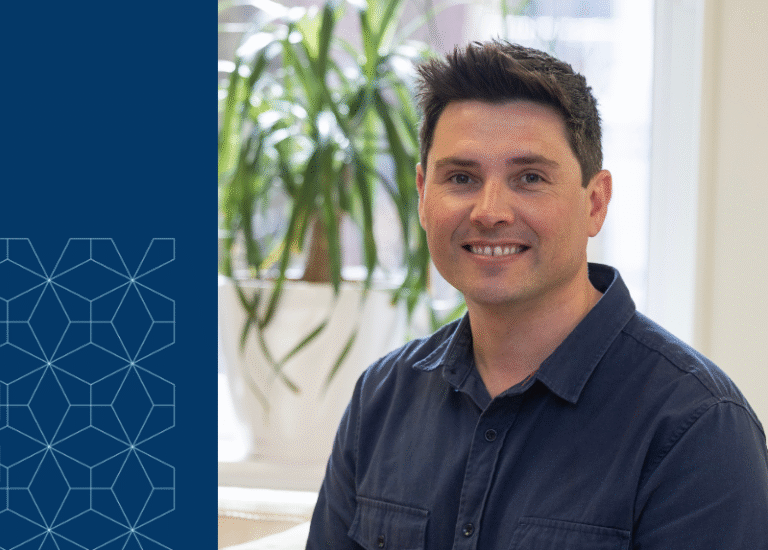 Smiling man in a navy shirt sitting indoors with a plant in the background and a geometric blue design on the left side of the image.