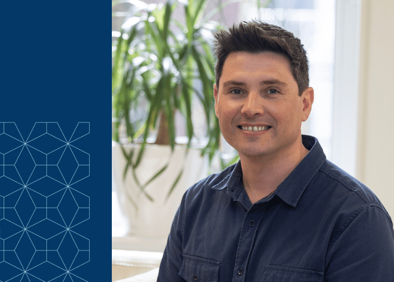 Smiling man in a navy shirt sitting indoors with a plant in the background and a geometric blue design on the left side of the image.